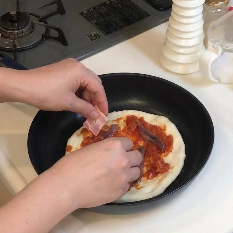 Hands placing rectangular slices of raw bacon onto a pizza already topped with tomato sauce and anchovies.