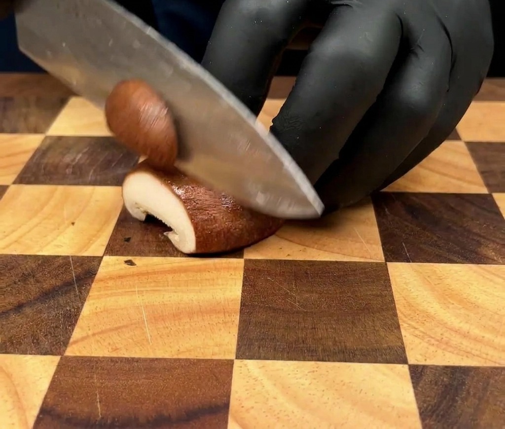 A close-up shot of a person wearing black gloves slicing a king oyster mushroom into thin sections on a wooden checkered cutting board.
