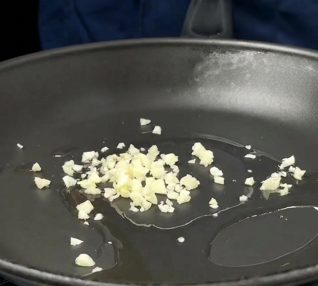 Minced garlic and chopped red onions sizzling in a thin layer of oil at the bottom of a dark frying pan.