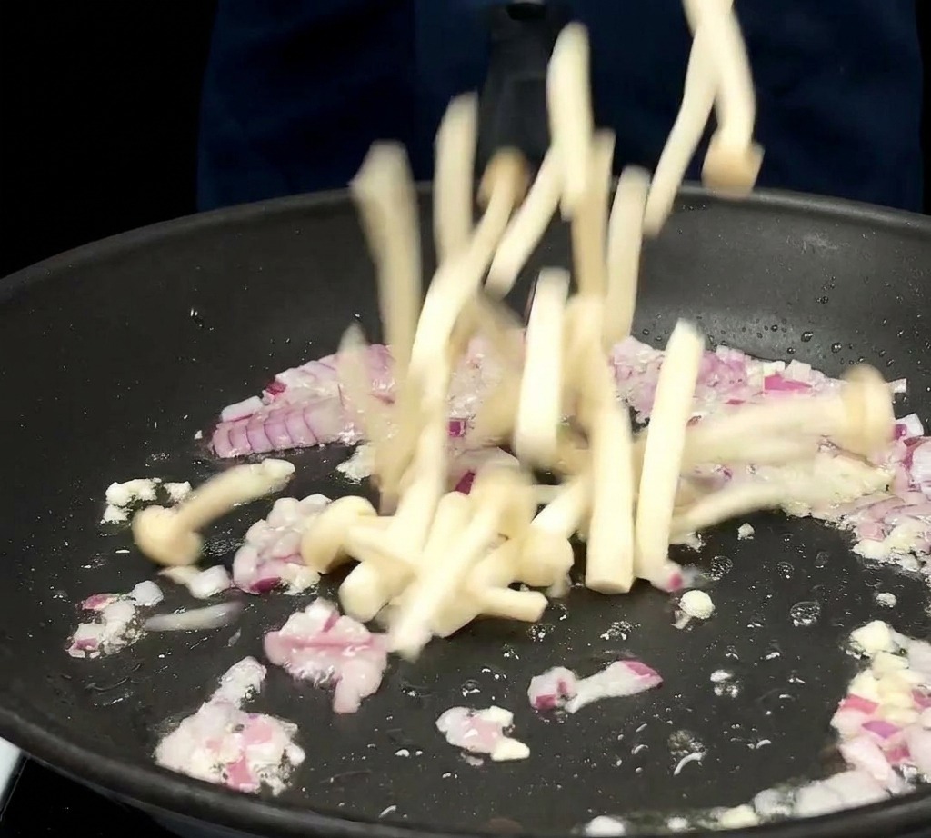 A bunch of long white jade mushrooms being dropped into a frying pan containing sautéed red onions and garlic.
