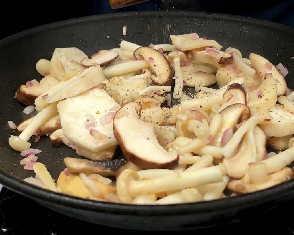 A person using a wooden pepper mill to grind black pepper over a variety of sautéed mushrooms and onions in a pan.