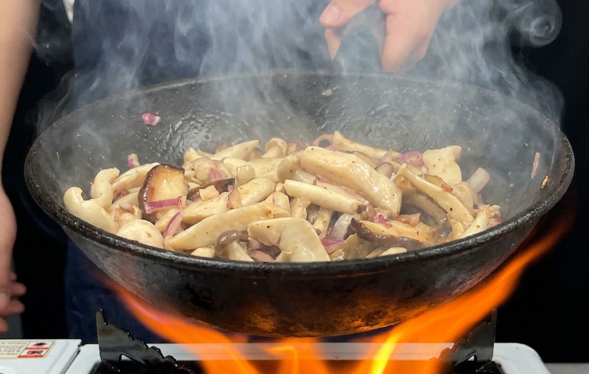 A frying pan filled with mushrooms being tossed over a large, bright orange gas flame with steam rising from the pan.