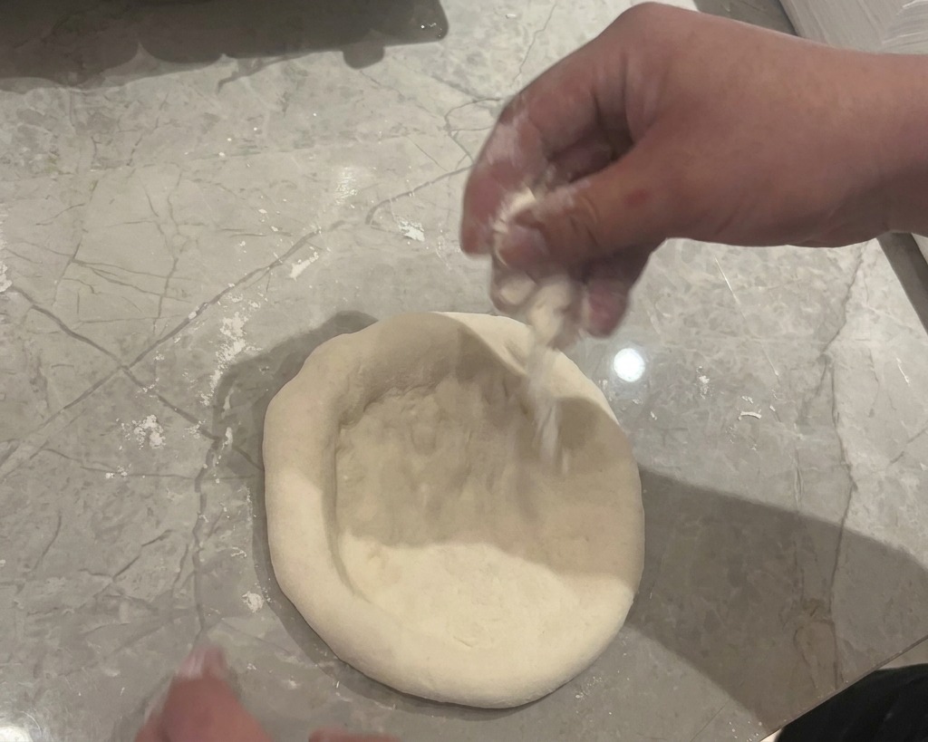 A pair of hands pressing and flattening a round ball of raw dough on a grey marble countertop dusted with white flour.
