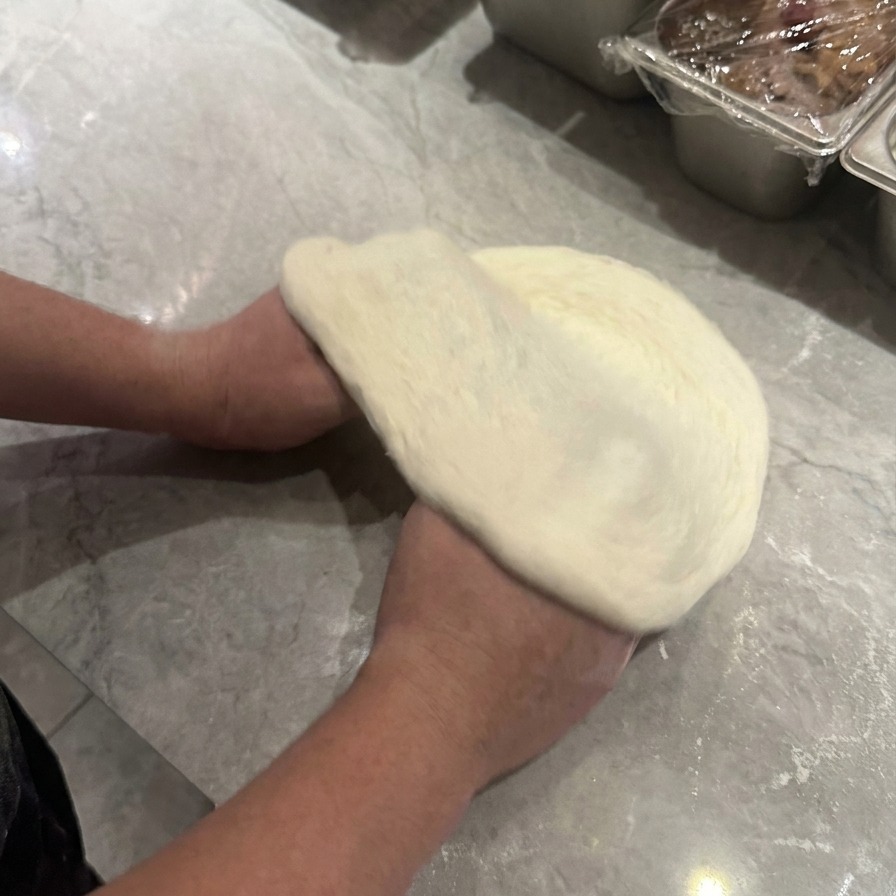 Two hands lifting and rotating raw pizza dough to stretch it into a circular shape with a visible outer rim.
