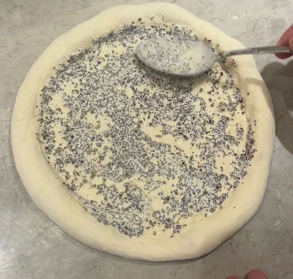 A metal spoon spreading a thick grey speckled truffle sauce over the center of a raw pizza base.