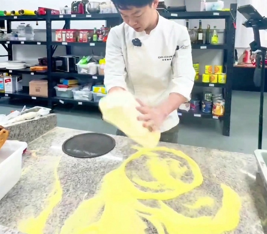 A chef in a white uniform vertically stretching a large pizza dough base in a professional kitchen.
