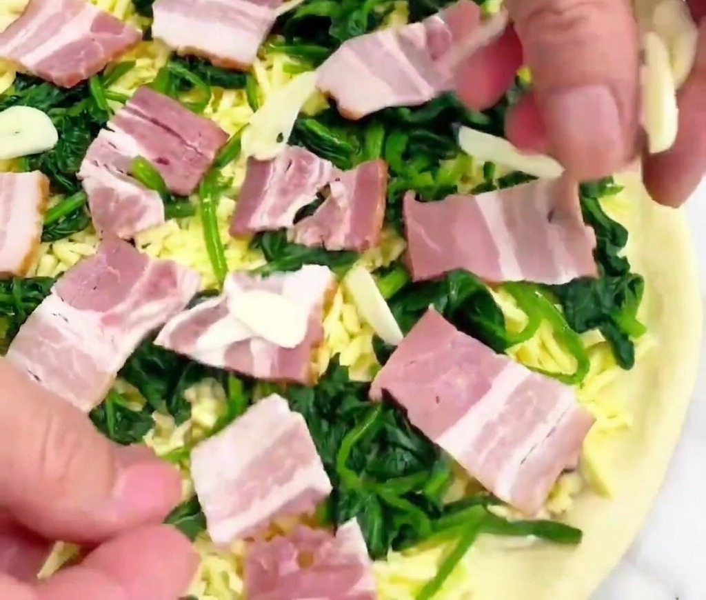 A hand dropping thin translucent garlic slices onto a raw pizza prepared with white sauce spinach bacon and cheese.