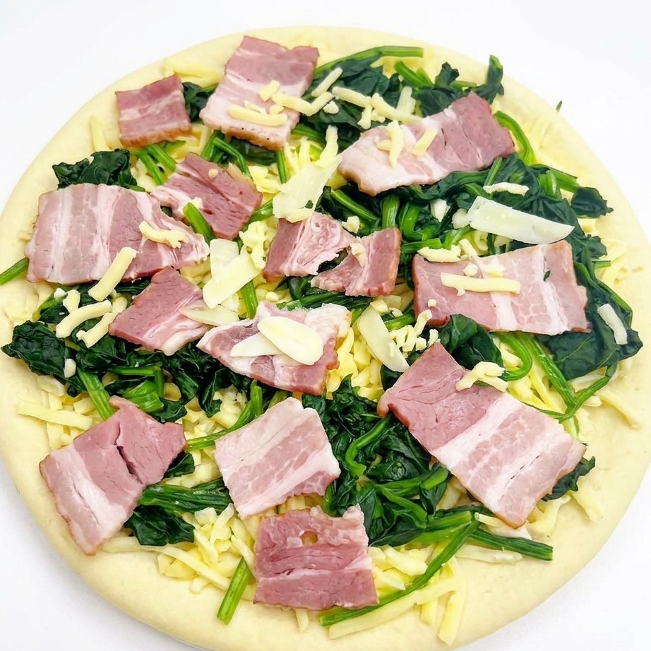 An overhead view of an assembled raw pizza with spinach, bacon, and garlic, topped with a light scattering of shredded mozzarella cheese.