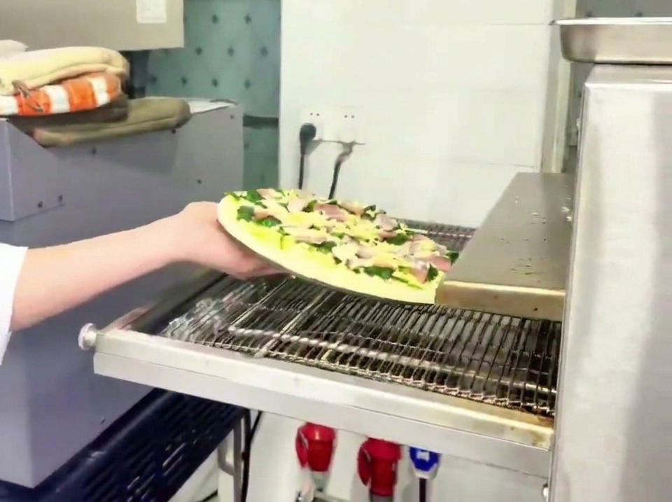 A worker placing the raw spinach and bacon pizza onto the moving metal belt of a professional pizza oven.