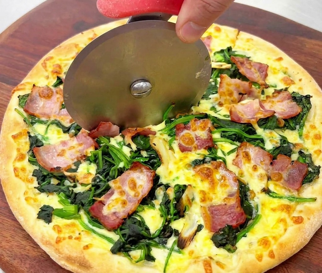 A close-up shot of a pizza wheel cutting through a golden-brown cooked pizza topped with wilted spinach and crispy bacon.