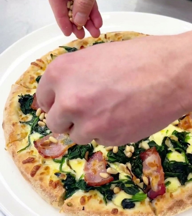 A hand scattering small toasted pine nuts over a freshly sliced spinach and bacon pizza on a white plate.