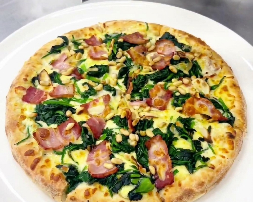A person using a grater to add fresh shavings of hard cheese to a cooked pizza garnished with spinach, bacon, and pine nuts.