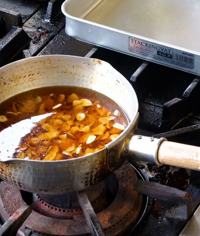 A variety of raw white garlic slices submerged in a mixture of liquid lard and sesame oil inside a hammered metal pot.