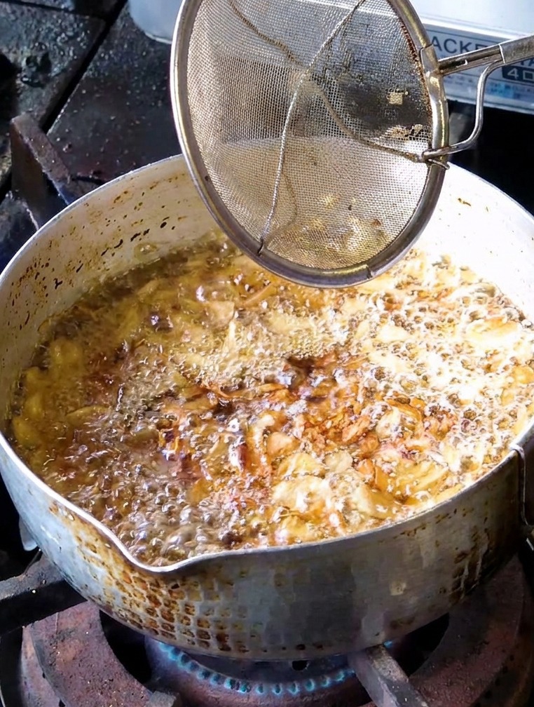 A mesh strainer being used to scoop out lightly fried golden brown garlic slices from a bubbling pot of oil.