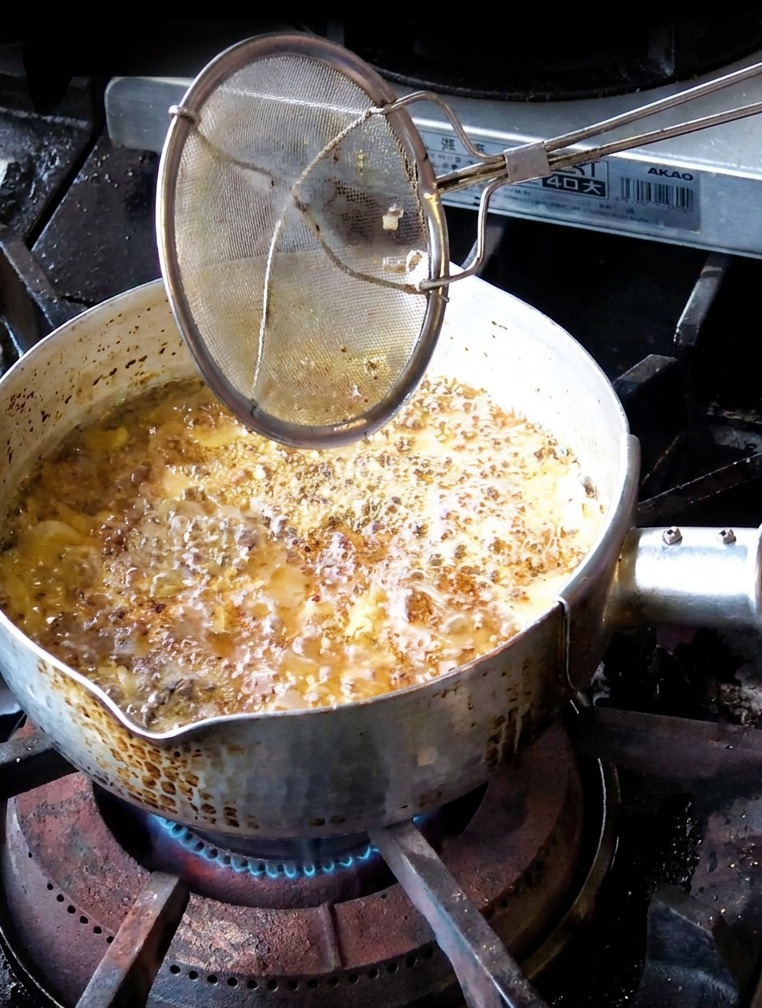 A hand using a mesh strainer to remove medium brown fried garlic slices from a pot of dark bubbling oil.