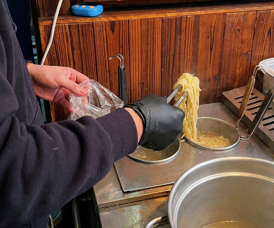 A person in black gloves using tongs to lift long, yellow ramen noodles from a boiling pot of water, preparing to place them into a bowl of broth.