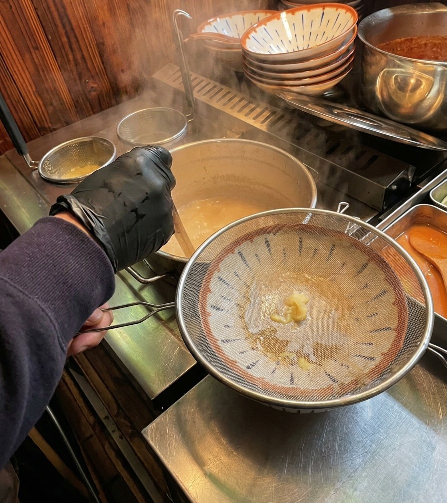 Steaming, creamy white bone broth being poured through a circular metal strainer into a decorated ramen bowl.