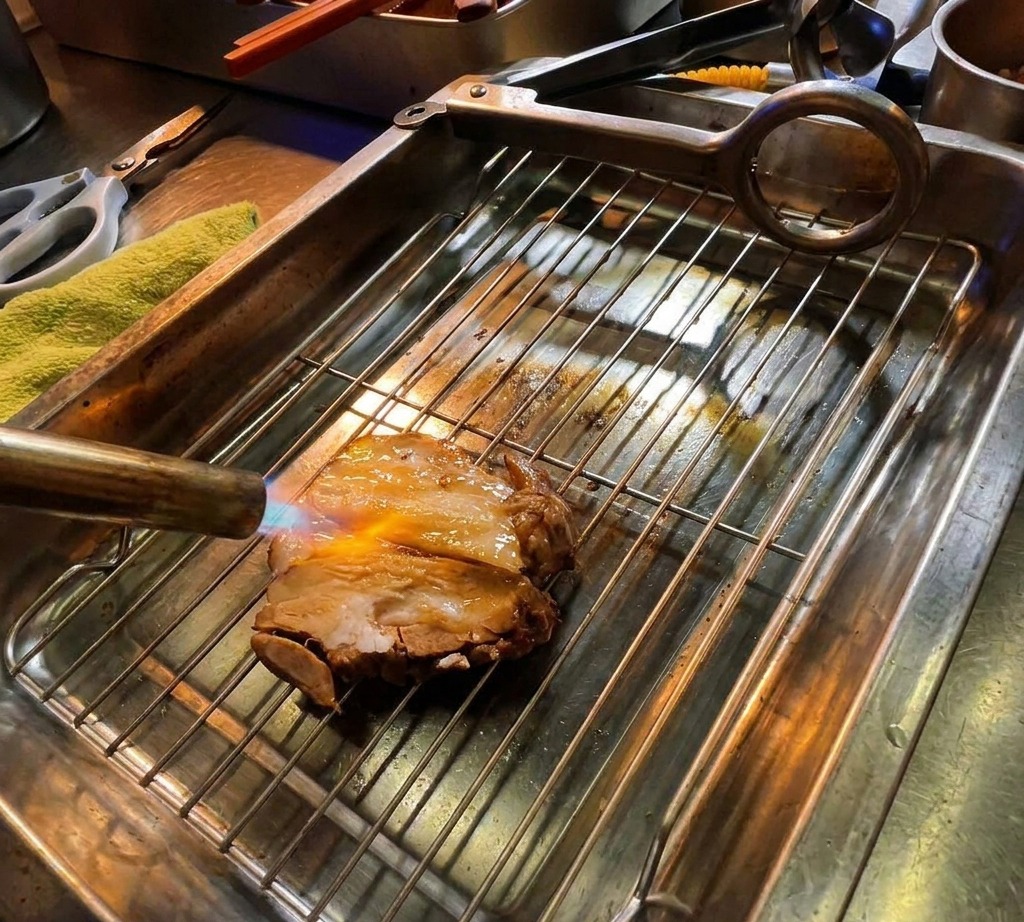 A kitchen blowtorch searing a thick slice of marinated pork chashu resting on a metal grate, with visible flames browning the meat.