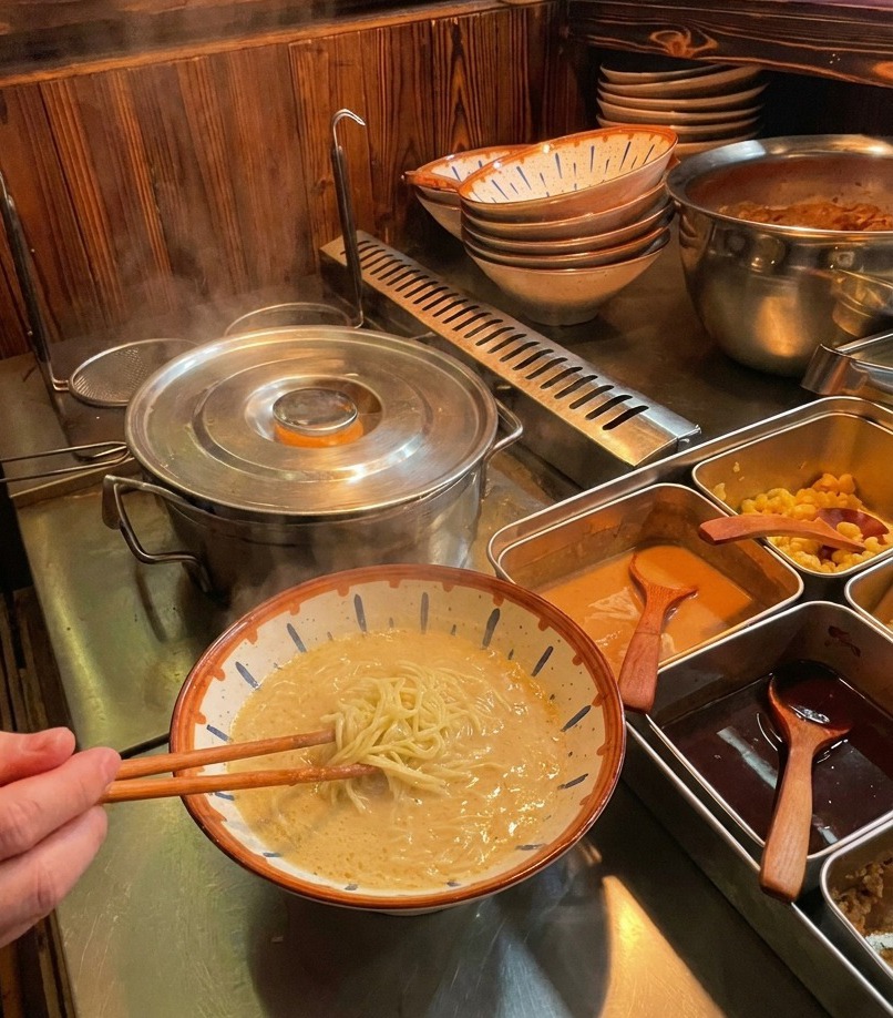 A person using wooden chopsticks to lift cooked ramen noodles and fold them into a bowl of creamy broth at a prep station.