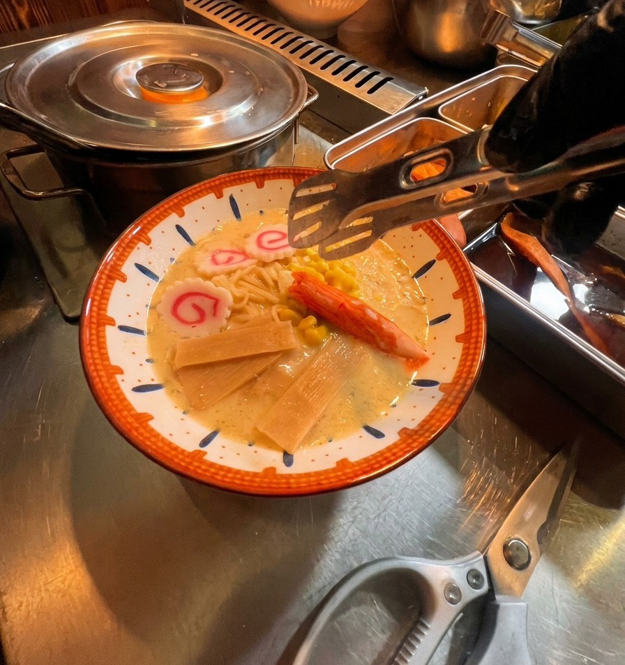 Tongs placing colorful toppings like pink fish cakes, a crab stick, and corn onto a bowl of ramen noodles and creamy broth.