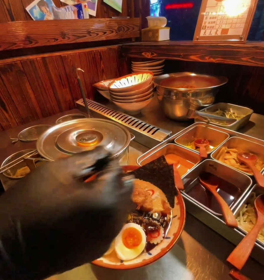 A black-gloved hand spooning dark, viscous garlic oil over a bowl of ramen filled with eggs, nori, and meat.