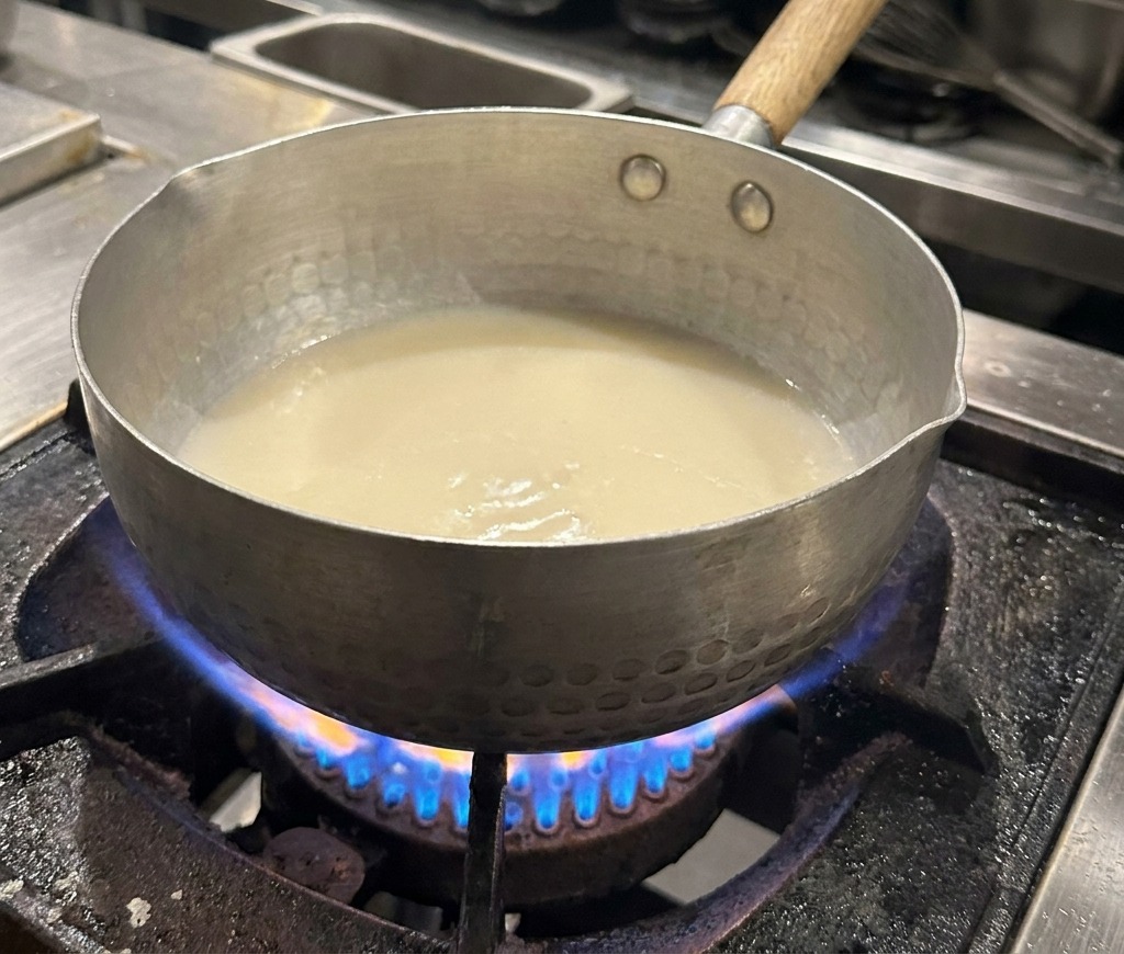 A close-up shot of a hammered metal pot on a stove filled with white broth, with subtle shimmering oil droplets appearing on the surface.