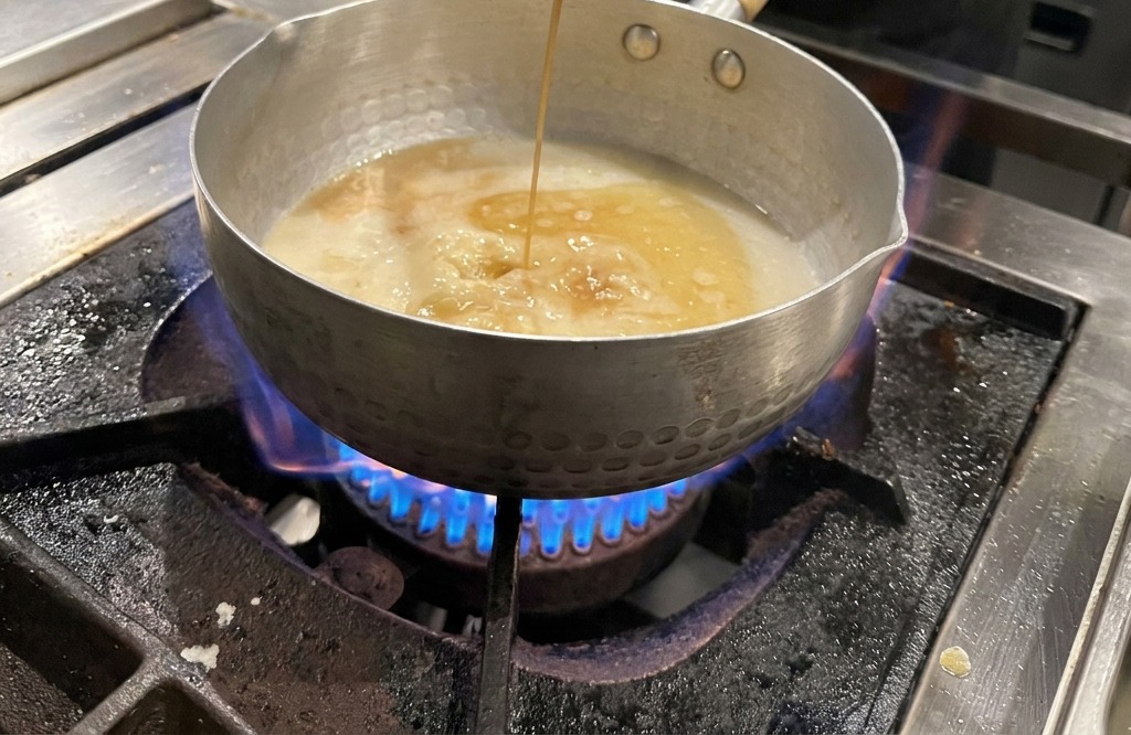 Dark soy sauce is poured from a metal ladle into a pot containing a light-colored broth over a lit gas burner.