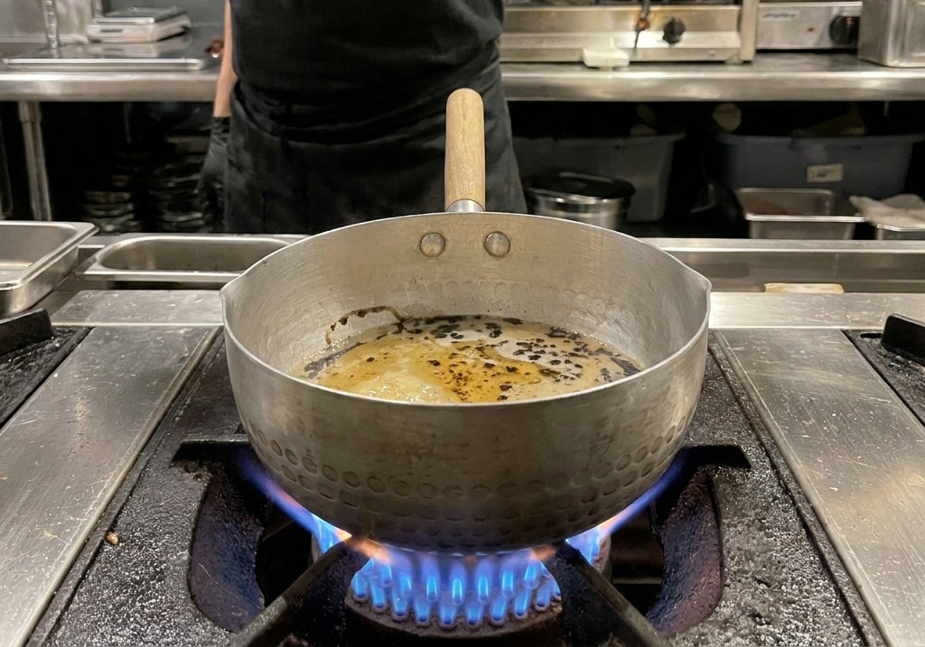 Dark black oil droplets and swirls float on top of a thickened, light-brown sauce inside a hammered metal pot.
