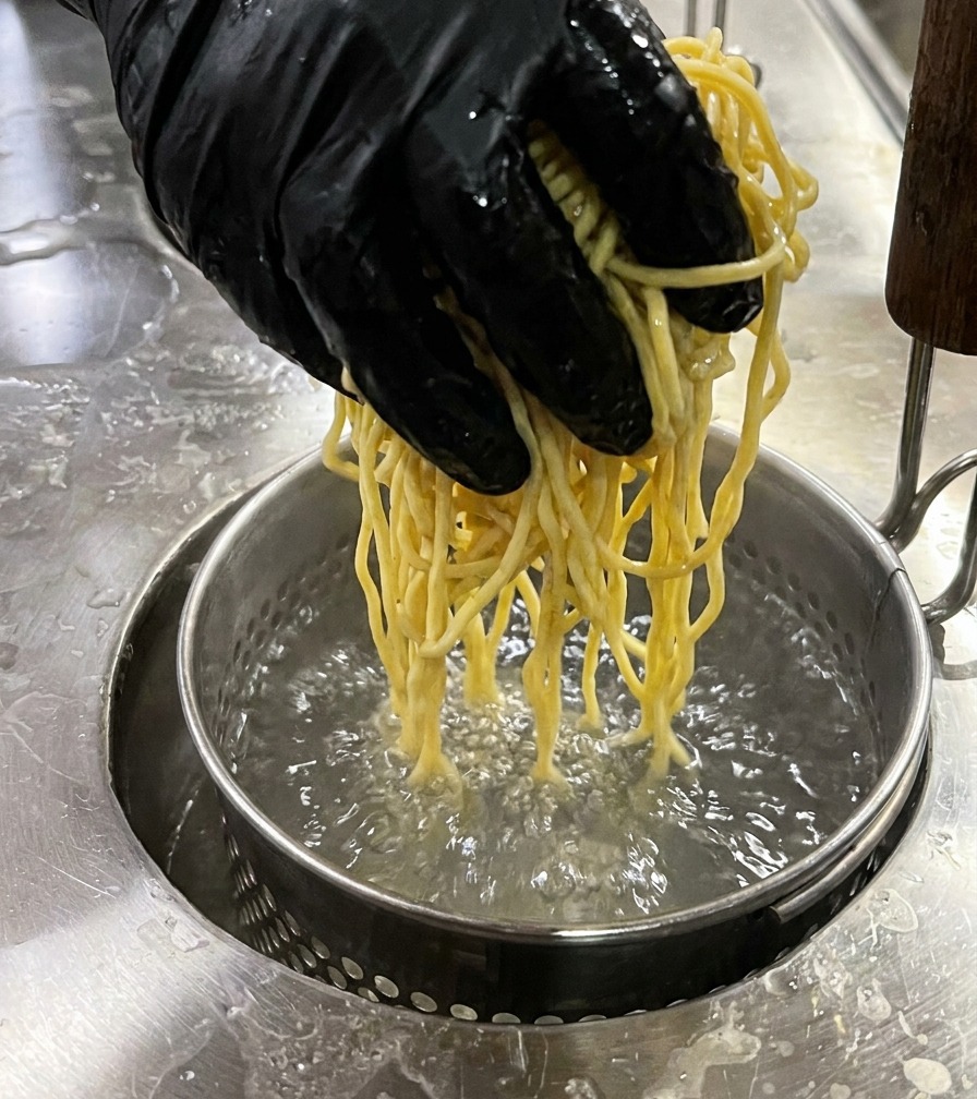 A hand in a black glove lowering a round metal noodle strainer filled with yellow noodles into boiling water.