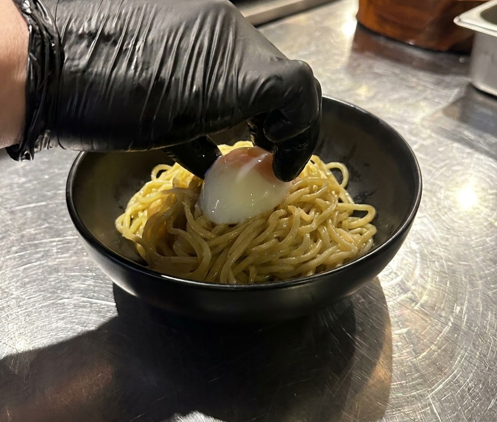A hand in a black glove gently placing a whole, soft-boiled onsen egg on top of a pile of ramen noodles in a black bowl.