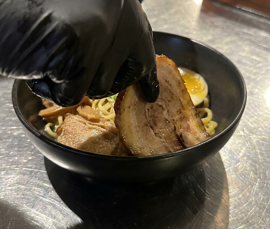 A gloved hand placing a thick slice of tender, seared chashu pork onto a bowl of noodles already garnished with a soft-boiled egg.