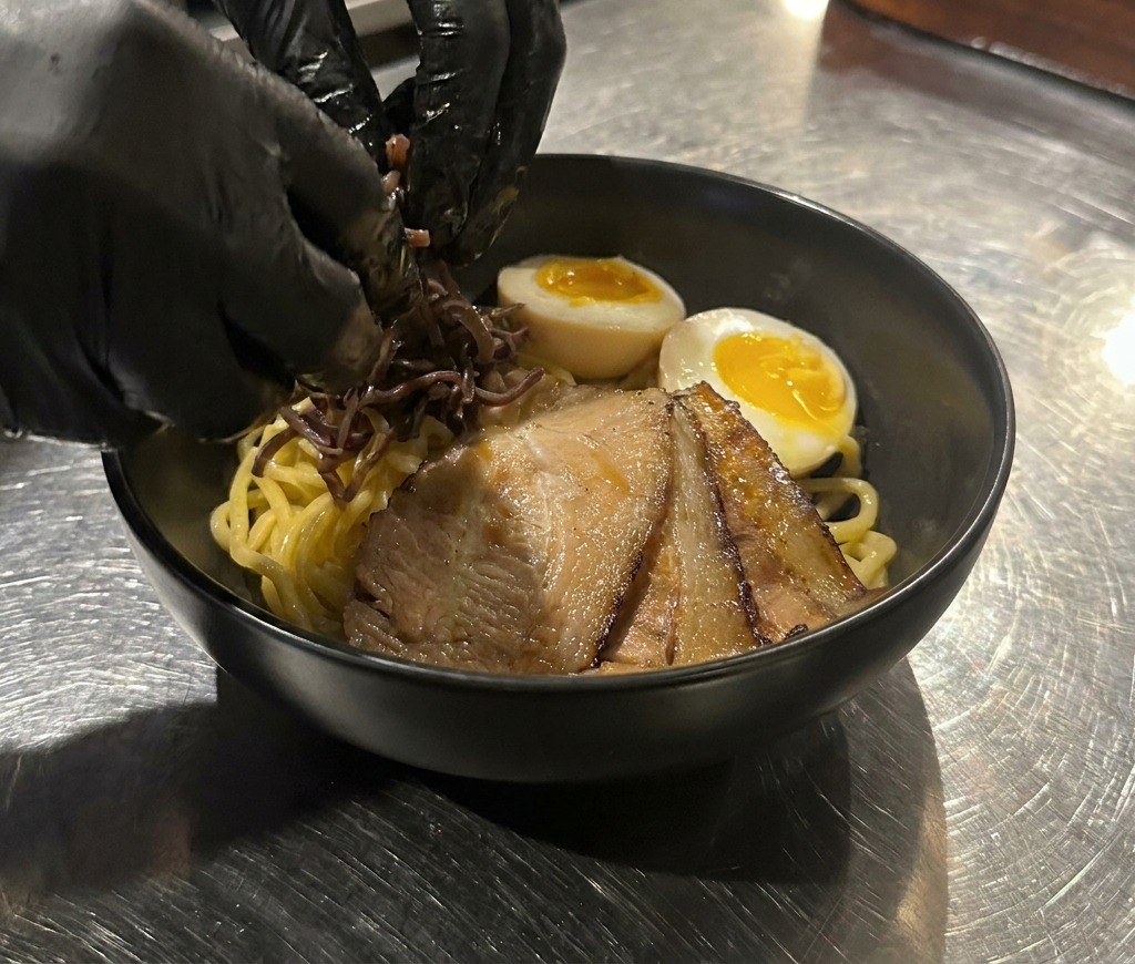 Gloved hands placing dark wood ear mushroom strips onto a bowl of noodles that already contains chashu and a soft-boiled egg.