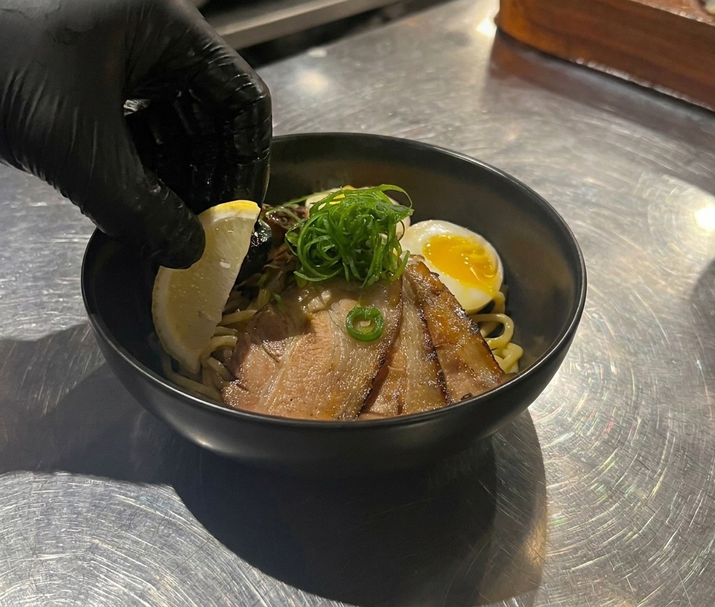 A hand placing a bright yellow lemon wedge into a black bowl filled with noodles, chashu pork, an egg, and green onion garnishes.