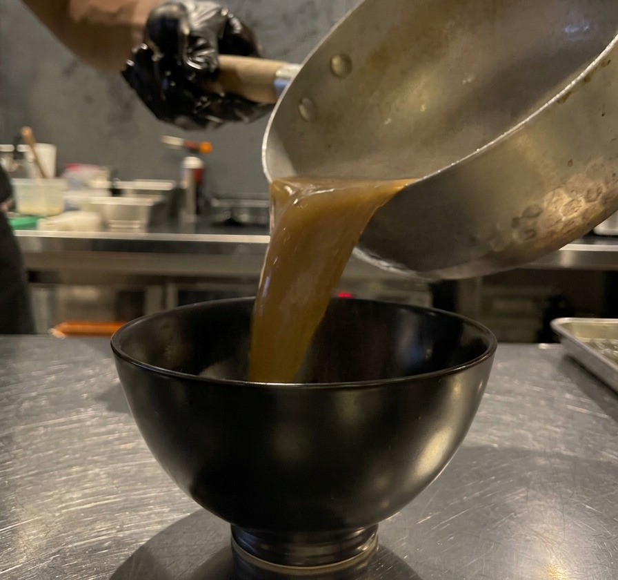 Thick, brown black garlic soup being poured from a silver cooking pot into a small black ceramic dipping bowl.