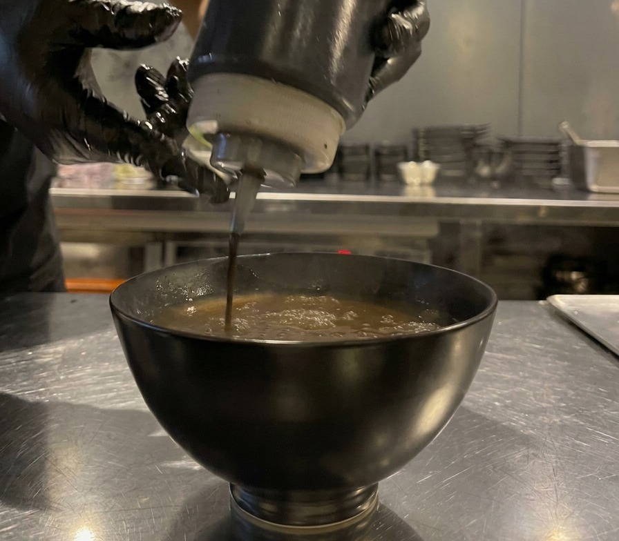 Dark black garlic oil being squeezed from a bottle into a bowl of brown dipping broth, creating dark droplets on the surface.