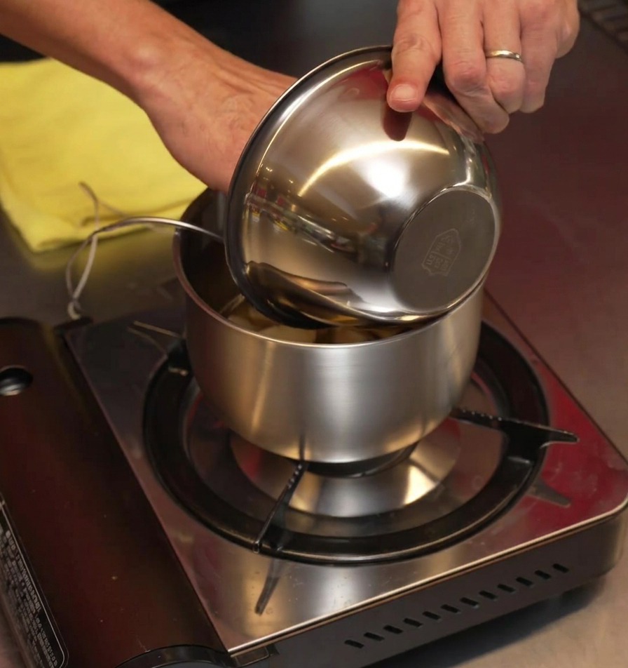 Fresh peeled garlic cloves being poured from a stainless steel bowl into a small pot on a portable gas stove.