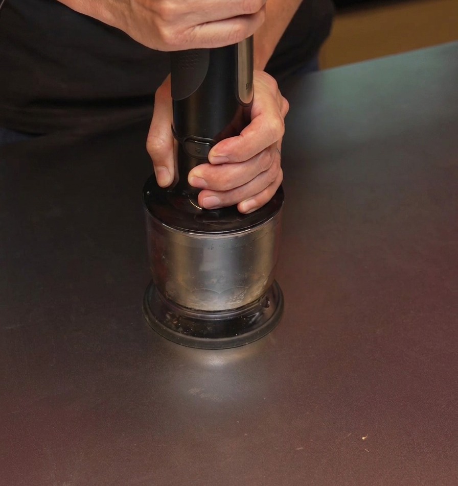 A person using a black hand blender to grind charred garlic inside a metal cup.