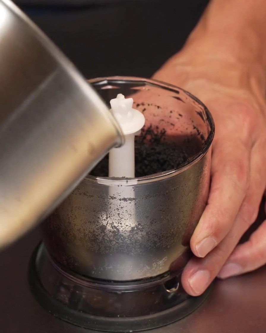 Darkened garlic infused oil being poured from a metal saucepan into a small blender jar containing black garlic paste.