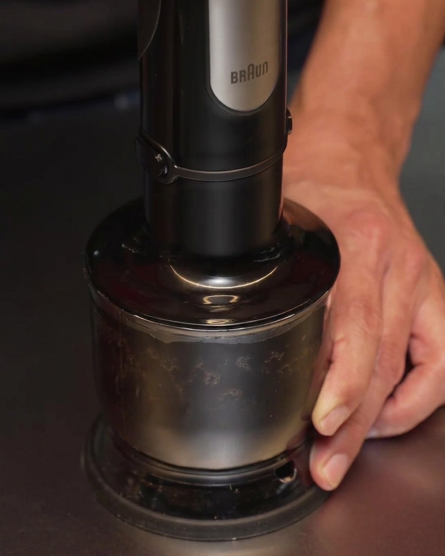 A hand blender being used to process blackened garlic and oil in a container, turning the mixture into a smooth, opaque black liquid.