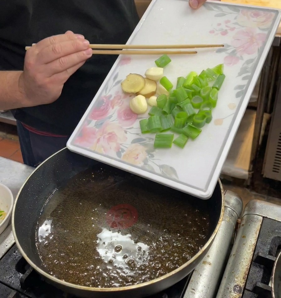 Hands holding a white floral board to slide chopped scallions, ginger, and garlic into a pan of hot oil.