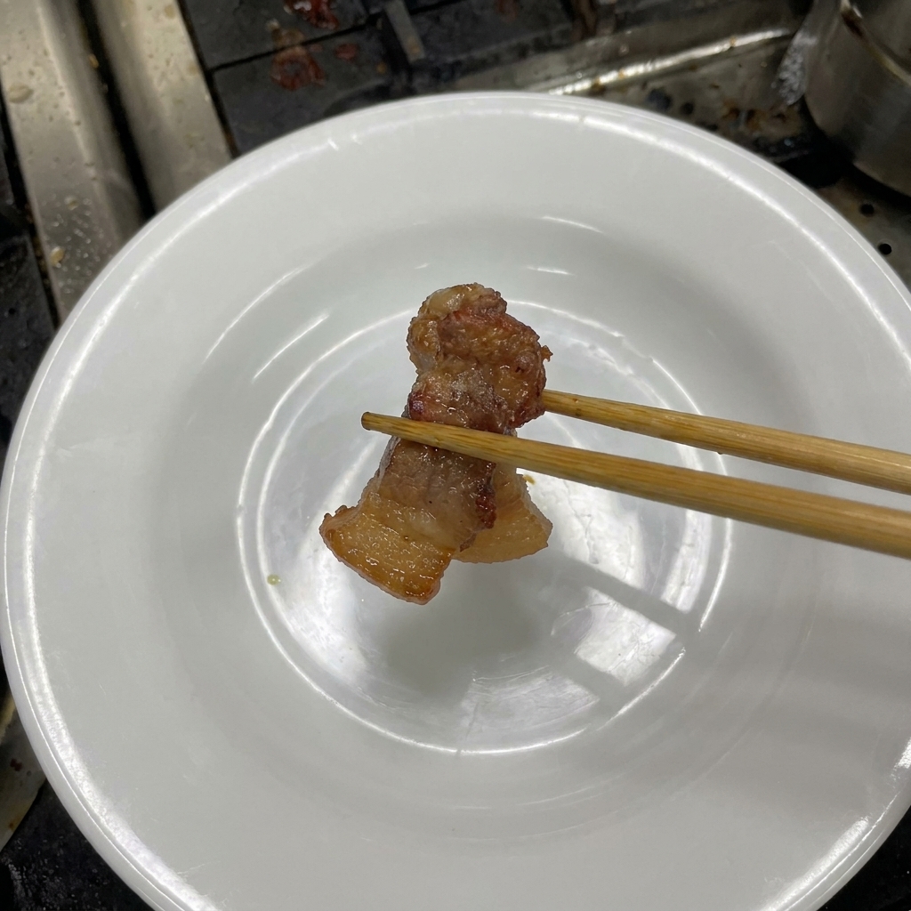 A pair of wooden chopsticks holding a piece of tender, dark brown braised pork belly over a white plate.