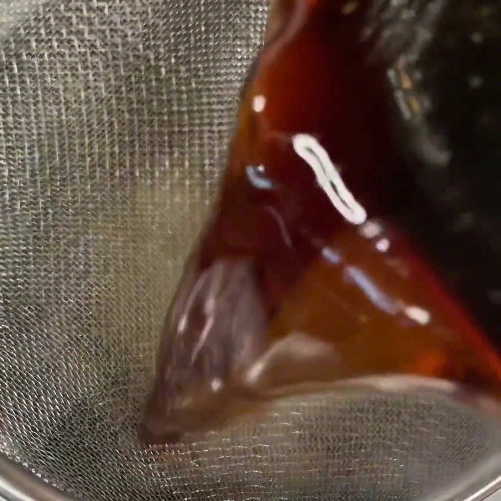 Close-up of a dark, translucent sauce being poured through a metal mesh strainer.