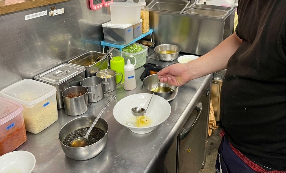 A ladle pouring clear, golden flavored oil into a white ceramic bowl on a stainless steel counter.