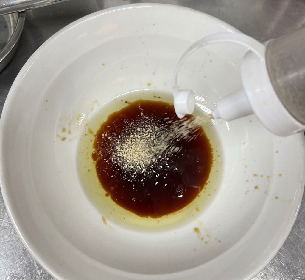 White sesame powder being sprinkled from a squeeze bottle into a white ceramic bowl containing a dark brown liquid sauce and clear flavored oil.