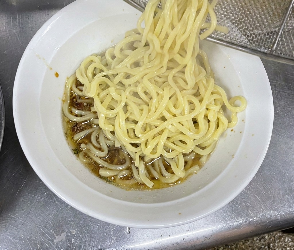 A bundle of thick, steaming noodles being dropped from a wire strainer into a prepared bowl.