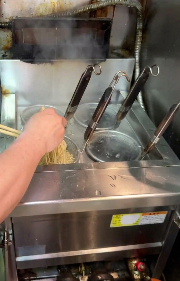 A cook lifting a mesh noodle strainer out of a boiler and shaking it over the water to drain the cooked ramen noodles.