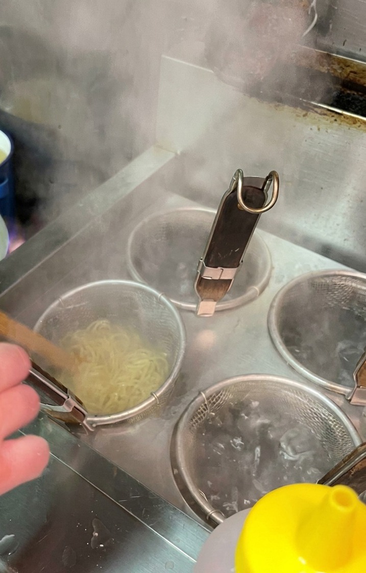 Noodles cooking inside a mesh strainer submerged in a pot of bubbling, boiling water in a professional kitchen.