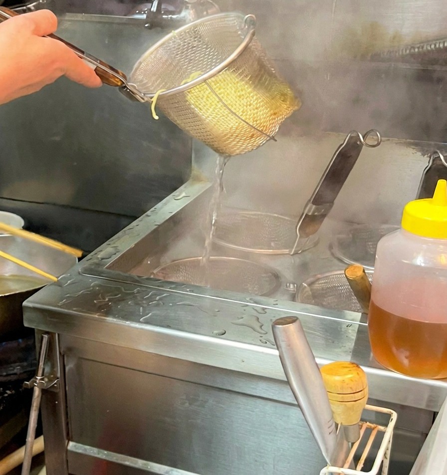 A hand holding a stainless steel noodle strainer over a bowl, releasing cooked ramen noodles into a dark, steaming broth.