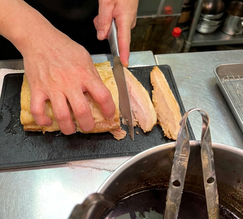 Close-up of a chef slicing a large piece of tender braised pork belly into thick slabs on a textured black cutting board.