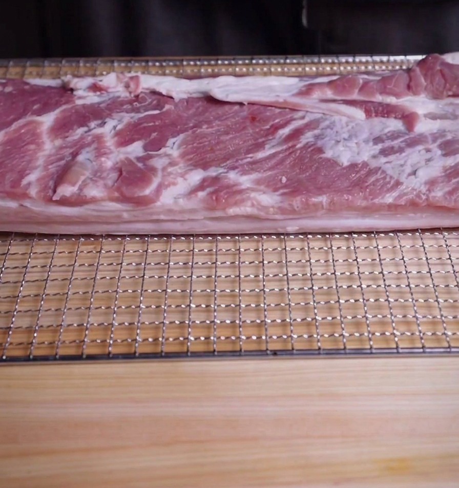 A large rectangular slab of raw pork belly resting on a metal wire rack on top of a wooden cutting board.